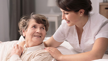 A young caregiver smiling and gently placing her hand on the shoulder of an elderly woman, who is sitting and looking up at her with a warm expression